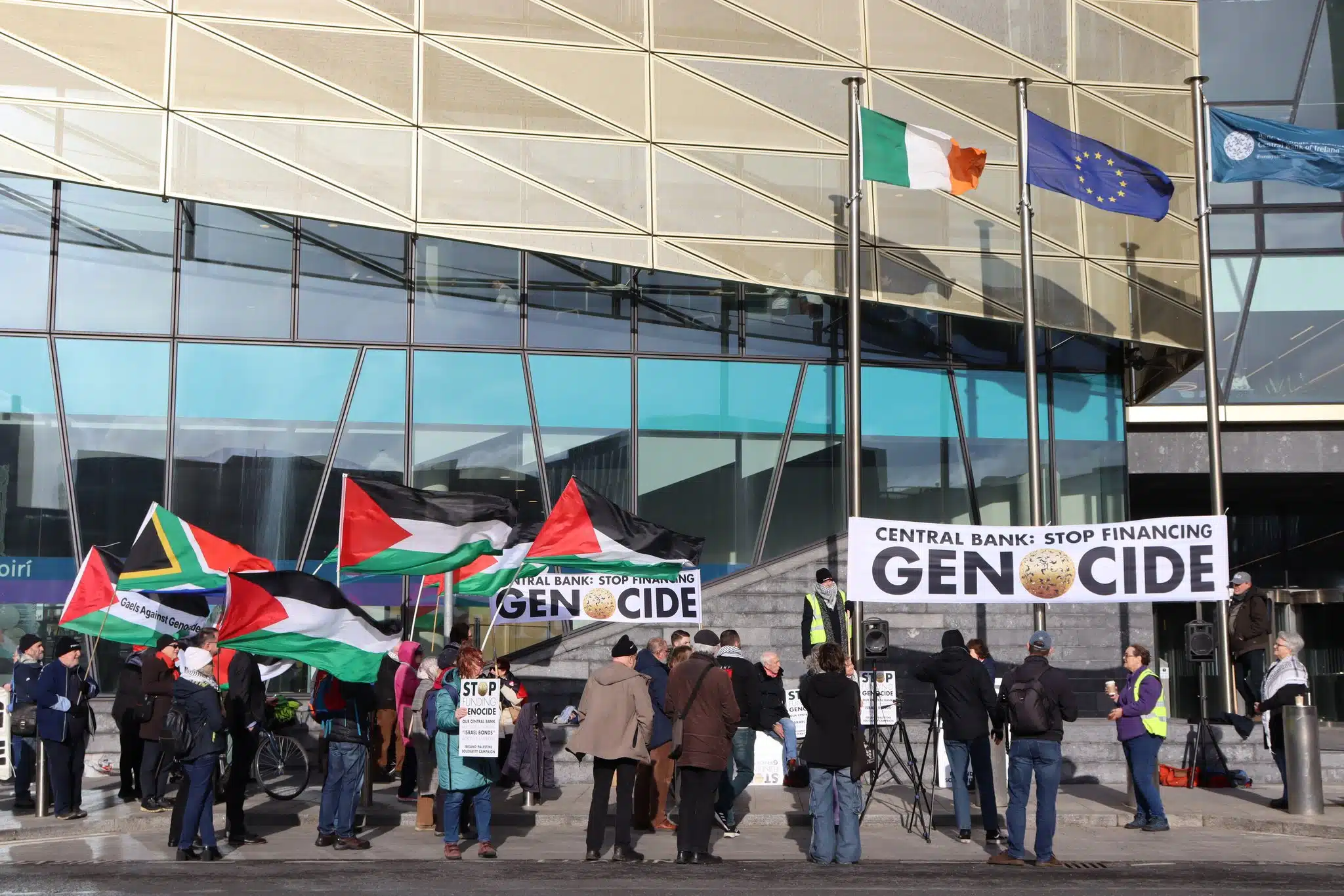 Protesters holding Palestinian flags and signs reading “Stop Financing Genocide” gather outside the Central Bank of Ireland, with Irish and EU flags flying above the entrance.