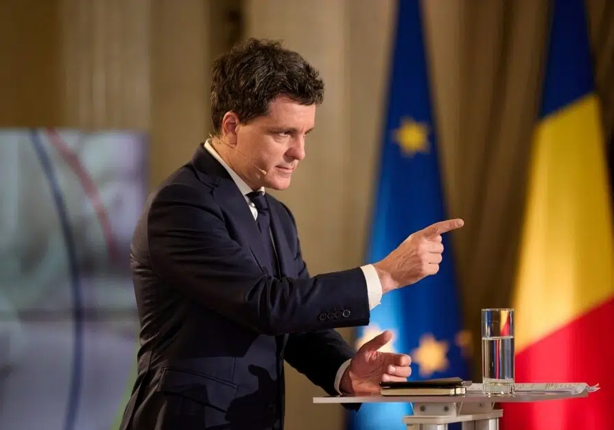 Nicușor Dan speaking emphatically at a podium, pointing with his finger, with EU and Romanian flags in the background