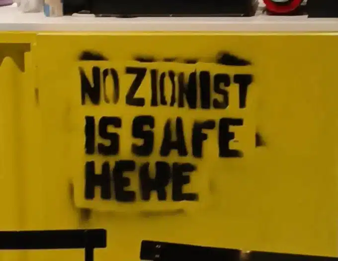 Spray-painted graffiti reading “No Zionist is safe here” on a yellow wall inside a restaurant.