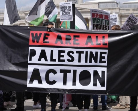 Pro-Palestinian protesters holding a large black banner reading "We Are All Palestine Action" with Palestinian flags in the background.