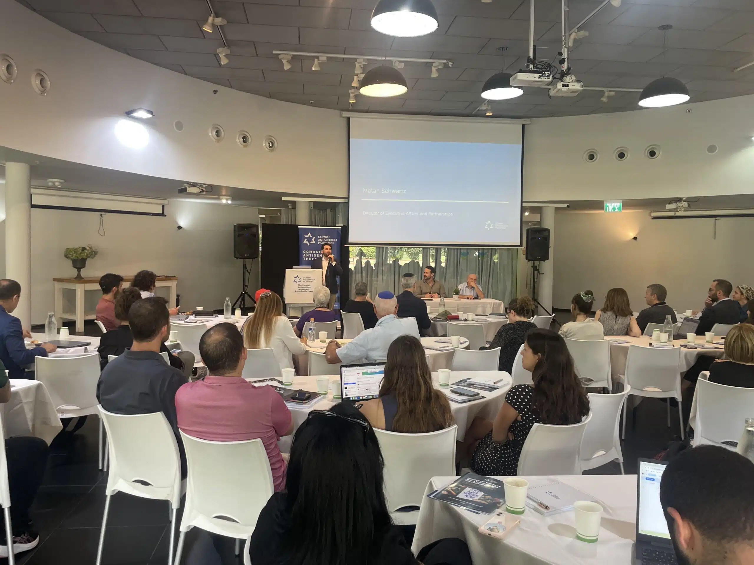 Participants seated around round tables listen to opening remarks at the CAM Partners Roundtable Forum in Tel Aviv.
