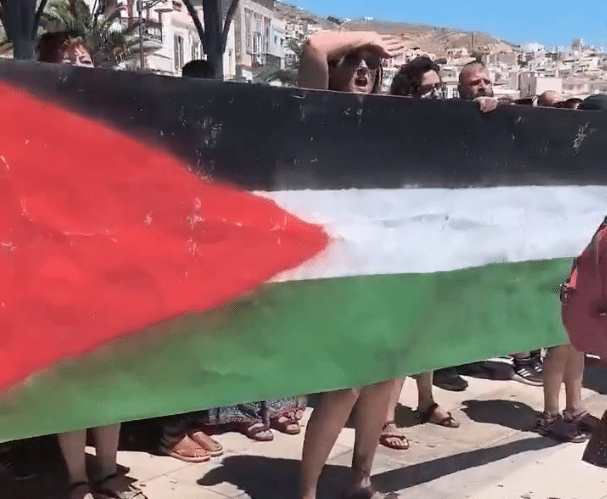 Pro-Palestinian protesters in Syros, Greece, holding a large Palestinian flag during a dockside demonstration.