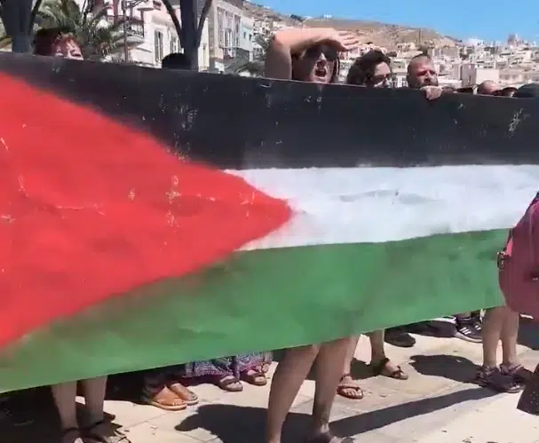 Pro-Palestinian protesters in Syros, Greece, holding a large Palestinian flag during a dockside demonstration.
