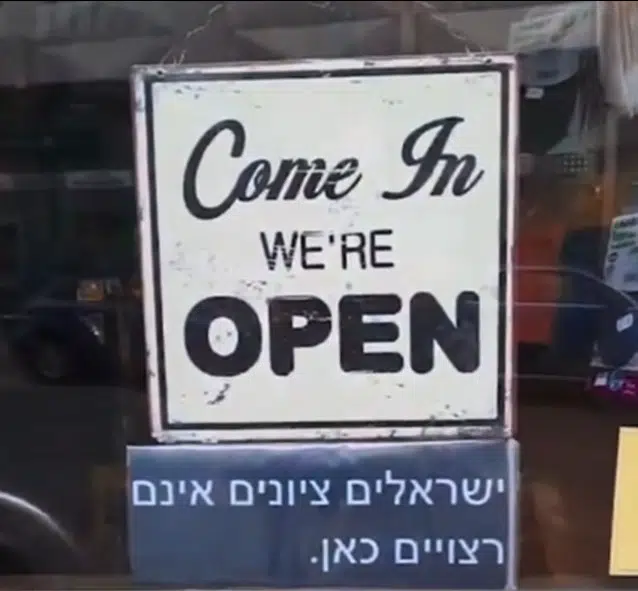 A storefront sign reads "Come In, We're Open," with a second sign below in Hebrew stating "Zionist Israelis are not welcome here."