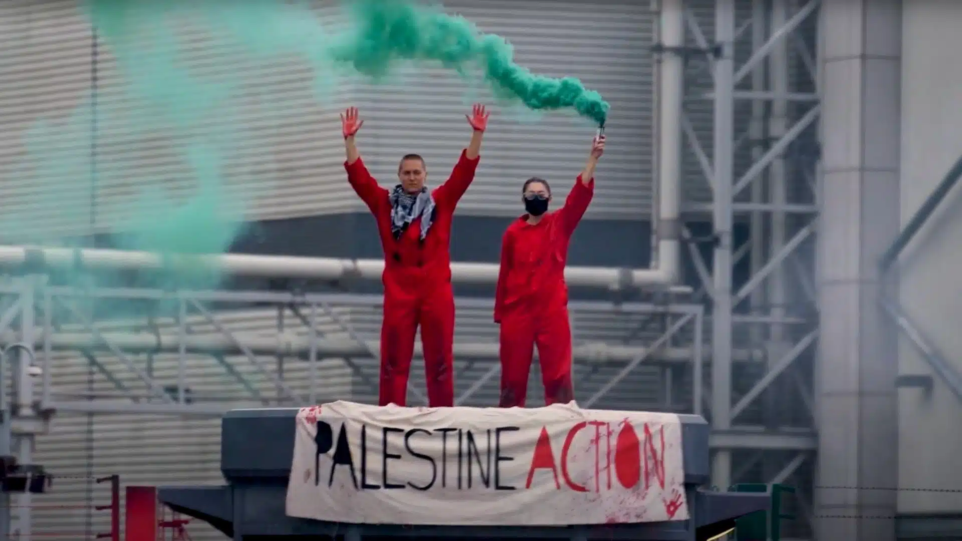 Two Palestine Action activists in red jumpsuits stand on a rooftop holding a green smoke flare above a banner reading "Palestine Action."