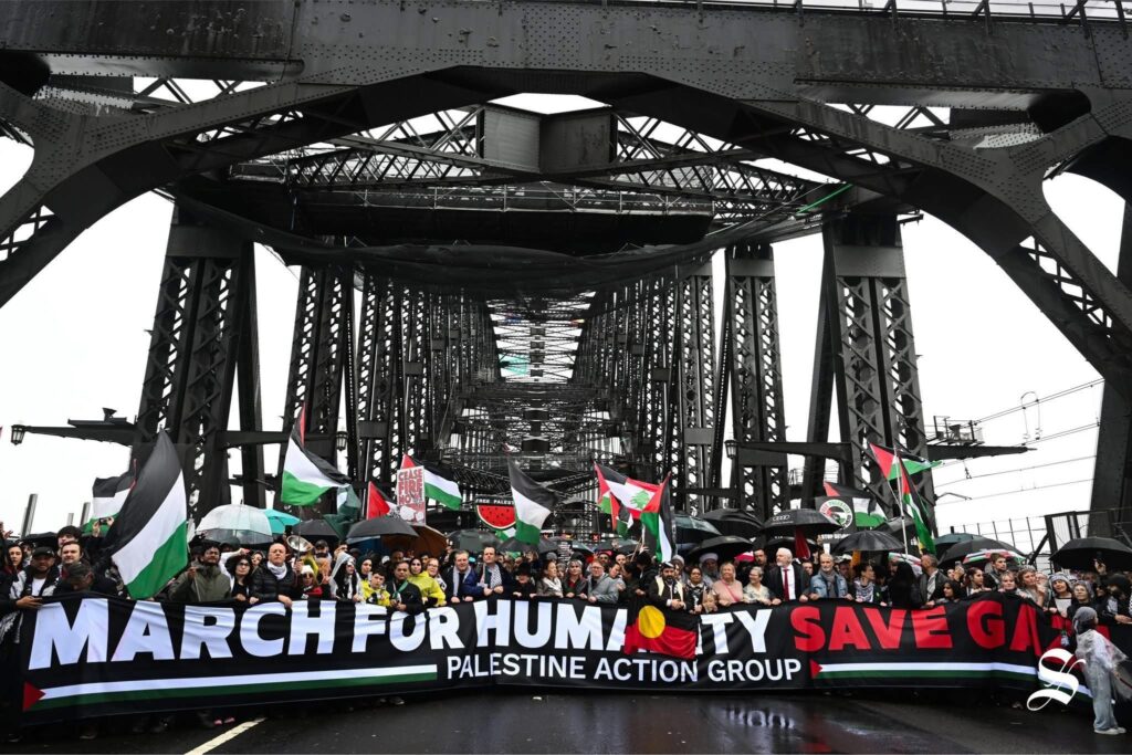 Pro-Palestinian protesters march across Sydney Harbour Bridge holding Palestinian flags and a banner reading “March for Humanity, Save Gaza – Palestine Action Group.”