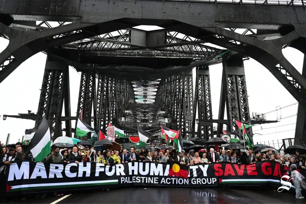 Pro-Palestinian protesters march across Sydney Harbour Bridge holding Palestinian flags and a banner reading “March for Humanity, Save Gaza – Palestine Action Group.”