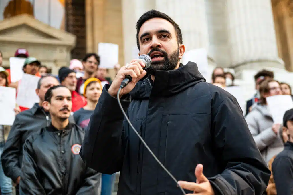 Zohran Mamdani speaks into a microphone at a rally, addressing a crowd of supporters gathered outside a government building.