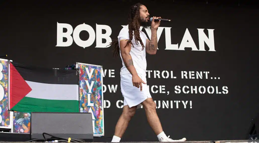 Bob Vylan’s frontman performing on stage, holding a microphone with a Palestinian flag displayed beside him.