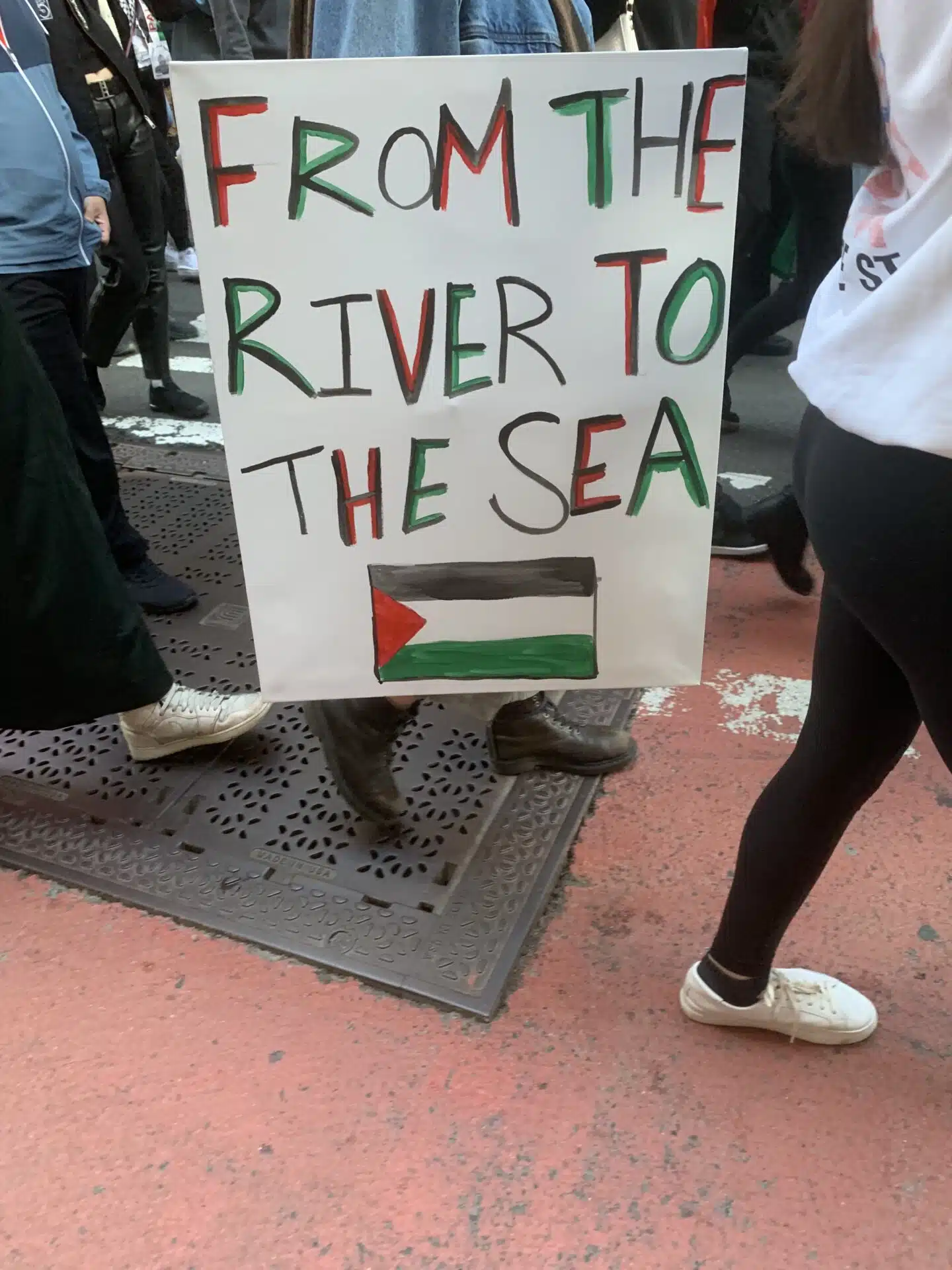 A protester holds a white sign reading “From the river to the sea” in red, black, and green letters, with a hand-drawn Palestinian flag at the bottom, during a street demonstration.