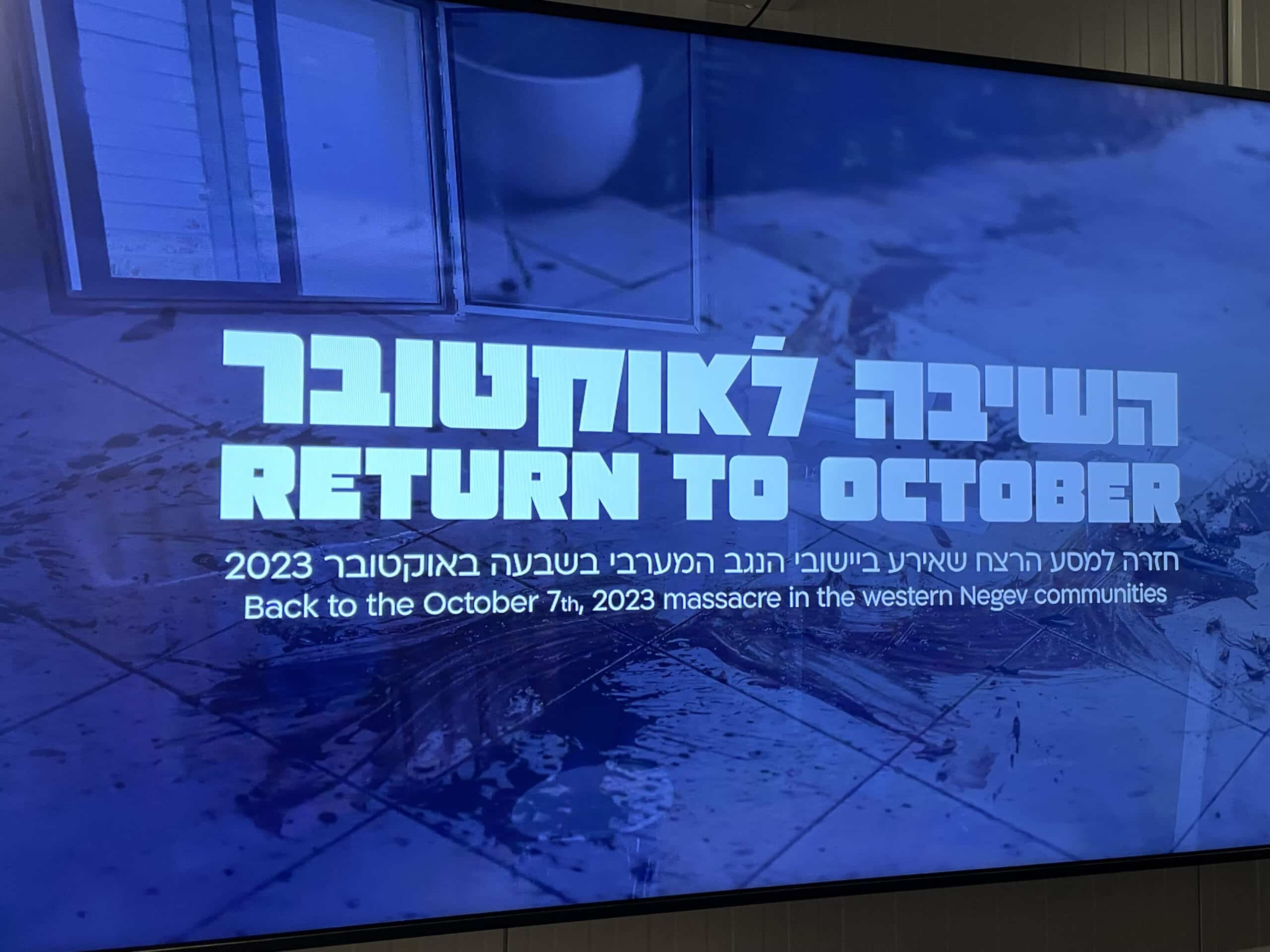 Digital display reading “Return to October” in Hebrew and English, shown against a background of bloodstained floor tiles — introducing an exhibit about the October 7, 2023 Hamas massacre in Israel’s western Negev communities.