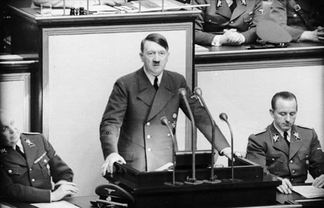 Adolf Hitler delivering a speech, surrounded by Nazi officials, in a historical archive photograph.