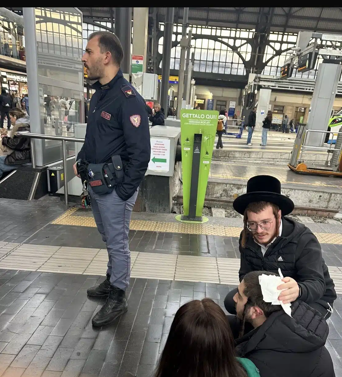 A police officer stands guard at Milan’s Central Station as Orthodox Jewish tourists tend to an injured companion after an antisemitic attack.