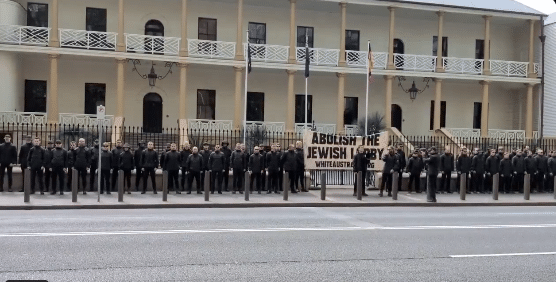Dozens of men dressed in black stand in formation outside a historic sandstone building in Sydney, Australia, holding a large antisemitic banner reading “Abolish the Jewish Lobby – White Australia.”