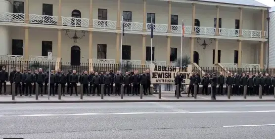 Dozens of men dressed in black stand in formation outside a historic sandstone building in Sydney, Australia, holding a large antisemitic banner reading “Abolish the Jewish Lobby – White Australia.”
