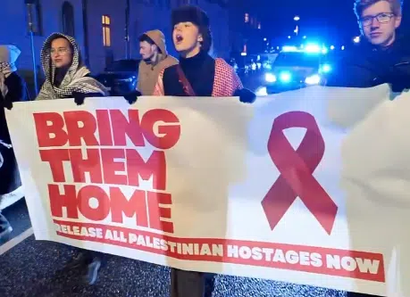 Anti-Israel activists in Stockholm holding a large red-ribbon banner reading “Bring Them Home – Release All Palestinian Hostages Now” during a nighttime demonstration outside the Israeli Embassy.