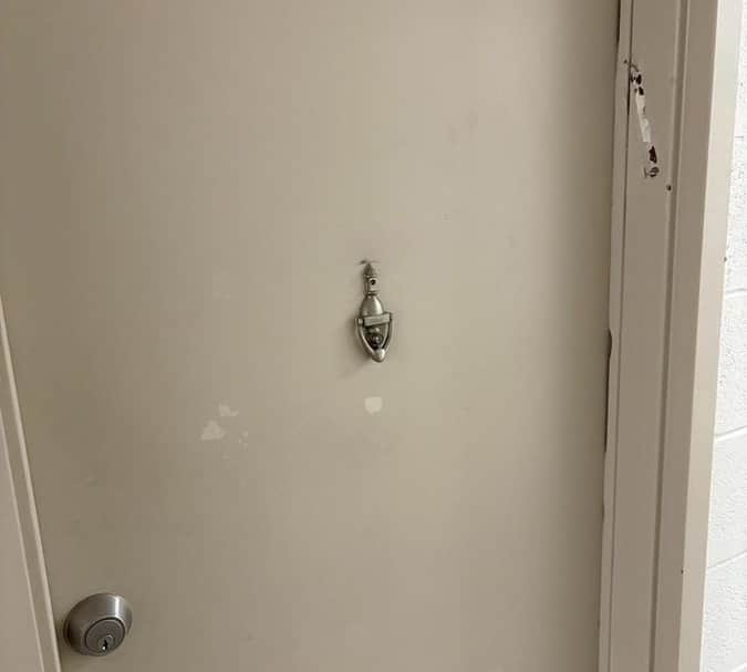 A beige apartment door in a North York senior housing building with clear damage where a mezuzah was forcibly removed from the right doorframe. The torn mounting strip and marks are still visible.