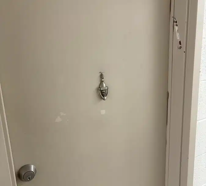 A beige apartment door in a North York senior housing building with clear damage where a mezuzah was forcibly removed from the right doorframe. The torn mounting strip and marks are still visible.