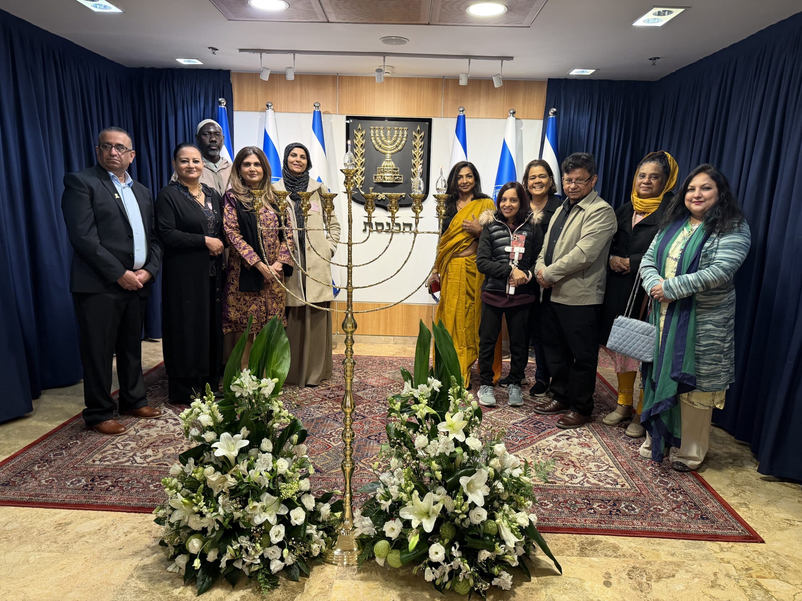 Members of the AMMWEC–CAM delegation stand beside a menorah inside the Israeli Knesset, with Israeli flags and the state emblem in the background.