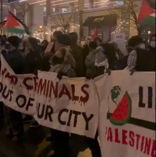 A crowd of anti-Israel protesters at night holding banners, including one reading “War criminals out of our city” and another with a watermelon symbol and “Free Palestine,” while waving Palestinian flags.