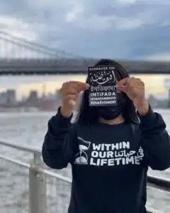 Person wearing a “Within Our Lifetime” sweatshirt holds a multilingual sticker reading “Globalize the Intifada” in English, Arabic, Bengali, Spanish, and French