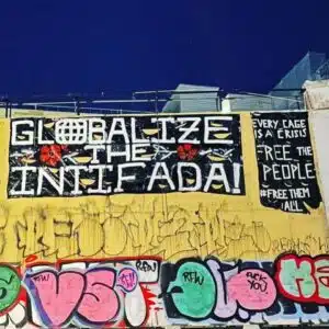 Graffiti on a wall reading “Globalize the Intifada,” painted above additional slogans calling to “Free the people” and “Free them all,” with layered street art beneath.