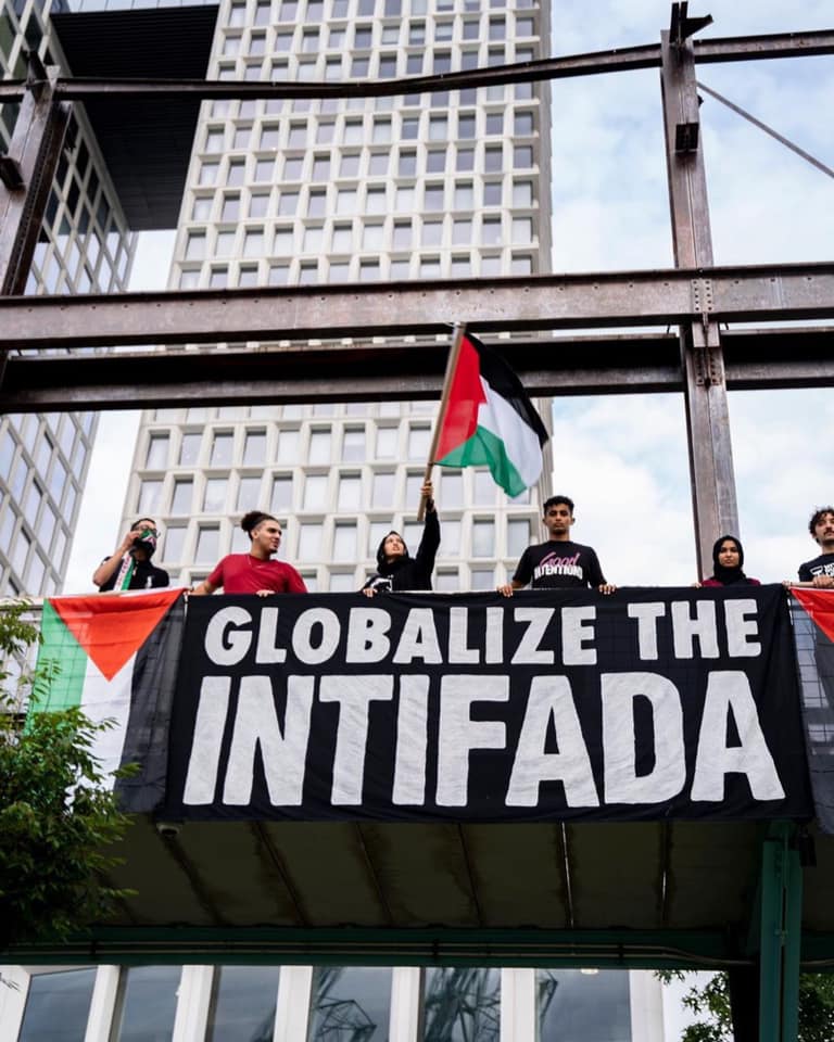 Pro-Palestinian demonstrators hang a banner reading “Globalize the Intifada” from a structure during a public protest, with Palestinian flags visible.