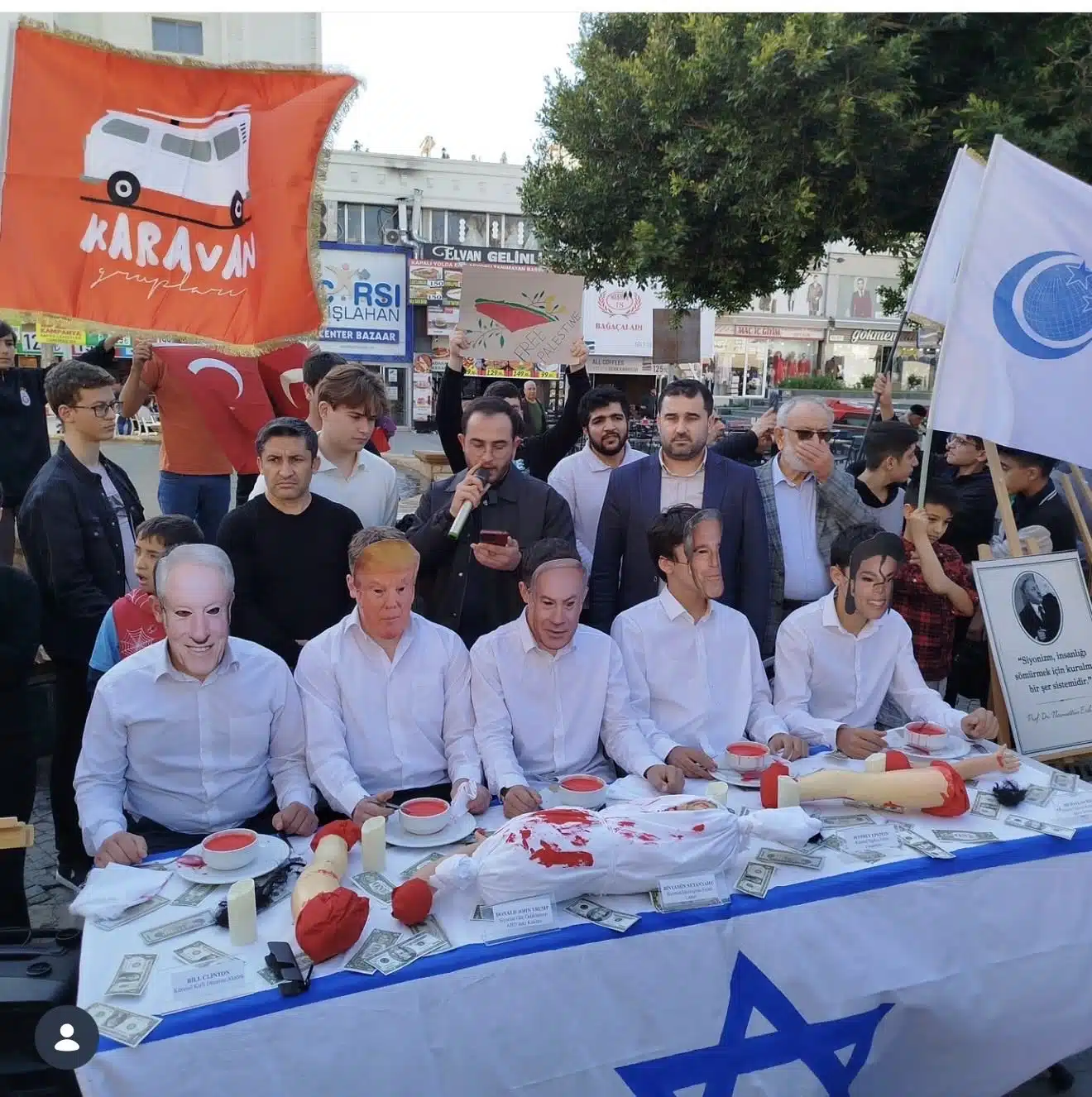 Public demonstration in Antalya, Turkey, showing masked figures of world leaders seated at a table staged with fake blood, childlike dolls, dollar bills, and an Israeli flag, as participants stand behind holding flags and signs.