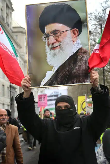 Masked demonstrator at an Al-Quds Day protest in London holds a large framed portrait of Ali Khamenei while Iranian flags are displayed behind him.