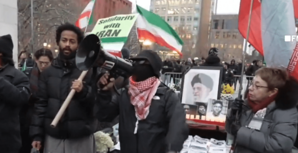 Masked demonstrator speaks through a megaphone at a pro-Khamenei vigil in Washington Square Park, New York City, with Iranian regime flags and a portrait of the late Iranian leader displayed behind him.