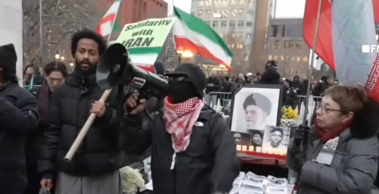 Masked demonstrator speaks through a megaphone at a pro-Khamenei vigil in Washington Square Park, New York City, with Iranian regime flags and a portrait of the late Iranian leader displayed behind him.