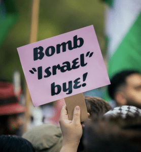 A protester holds a pink sign reading “Bomb ‘Israel’ bye!” above a crowd at a demonstration.