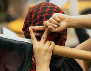 Close-up of a protester wearing a red-and-black keffiyeh covering their face, forming an inverted triangle hand gesture in front of their eye while holding a Palestinian flag