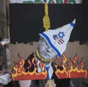 A protest sign depicting a caricature of a man wearing a white pointed hood with symbols including a Star of David and U.S. flag elements, with a noose around his neck and flames rising below.