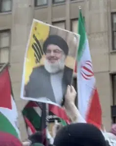 A protester holds up a poster of Hassan Nasrallah in a crowd, with Iranian and Palestinian flags visible in the background