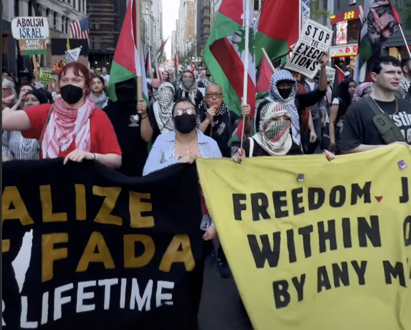 A large group of protesters marches through NYC carrying Palestinian flags, banners, and signs, including one reading “Abolish Israel.”