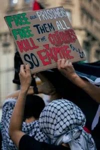 Protester holding a sign reading “Free our prisoners, free them all, kill the colonizers so empire falls” during a pro-Palestinian demonstration, with participants wearing keffiyehs in the background.
