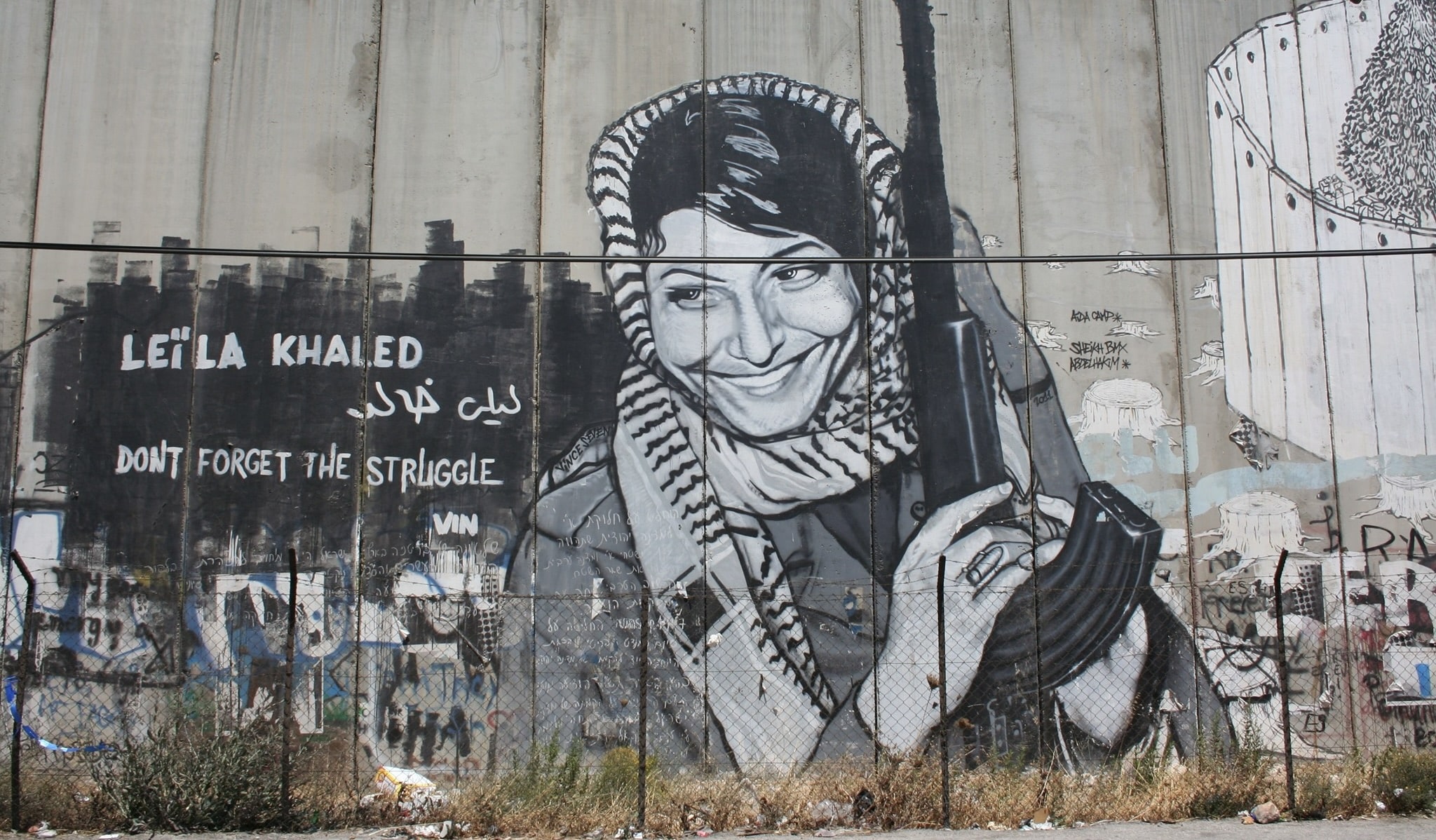 Graffiti mural of PFLP terrorist and airplane hijacker Leila Khaled holding a rifle on Israel’s security barrier, alongside the words “Don’t Forget the Struggle.”