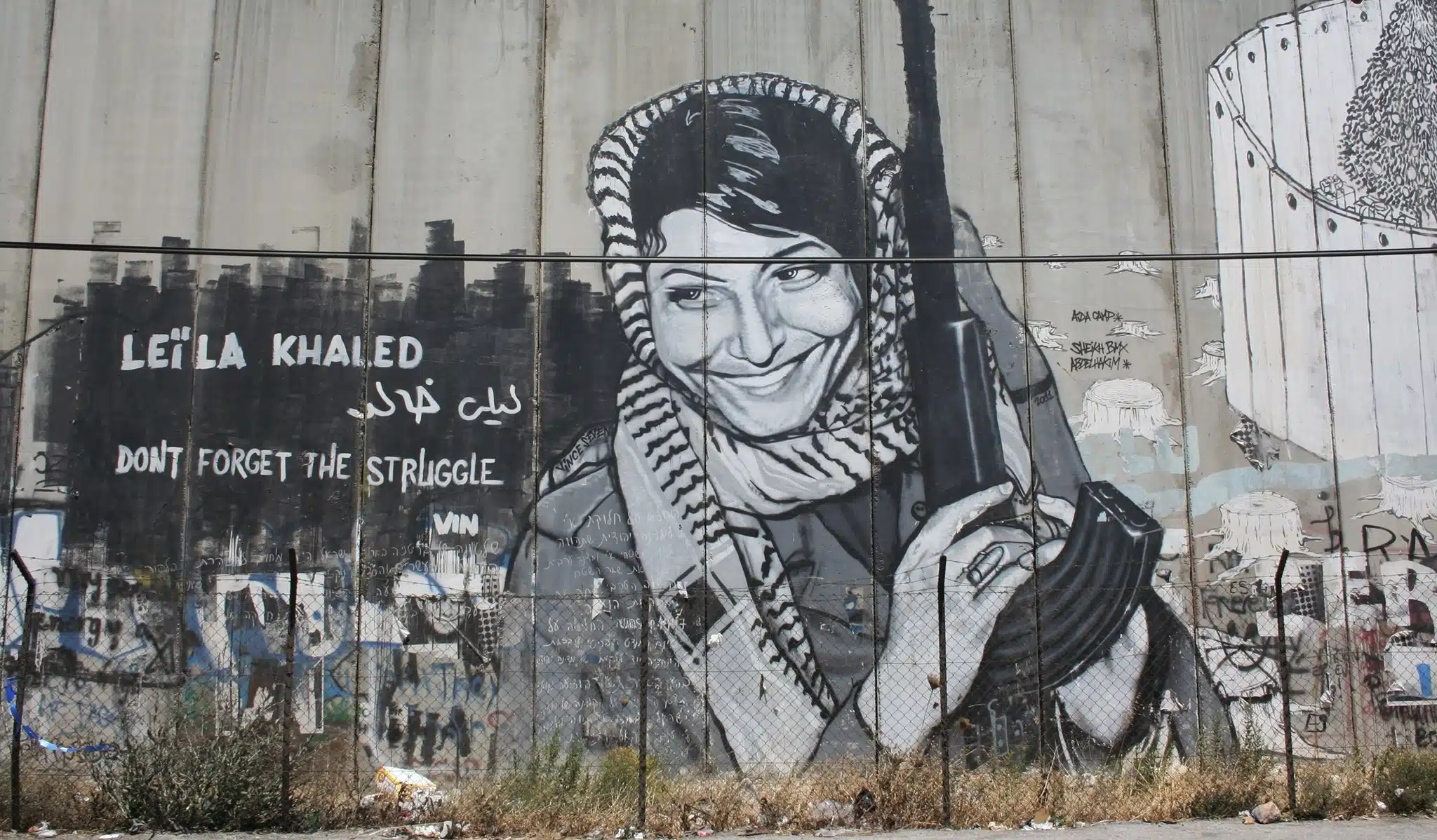 Graffiti mural of PFLP terrorist and airplane hijacker Leila Khaled holding a rifle on Israel’s security barrier, alongside the words “Don’t Forget the Struggle.”