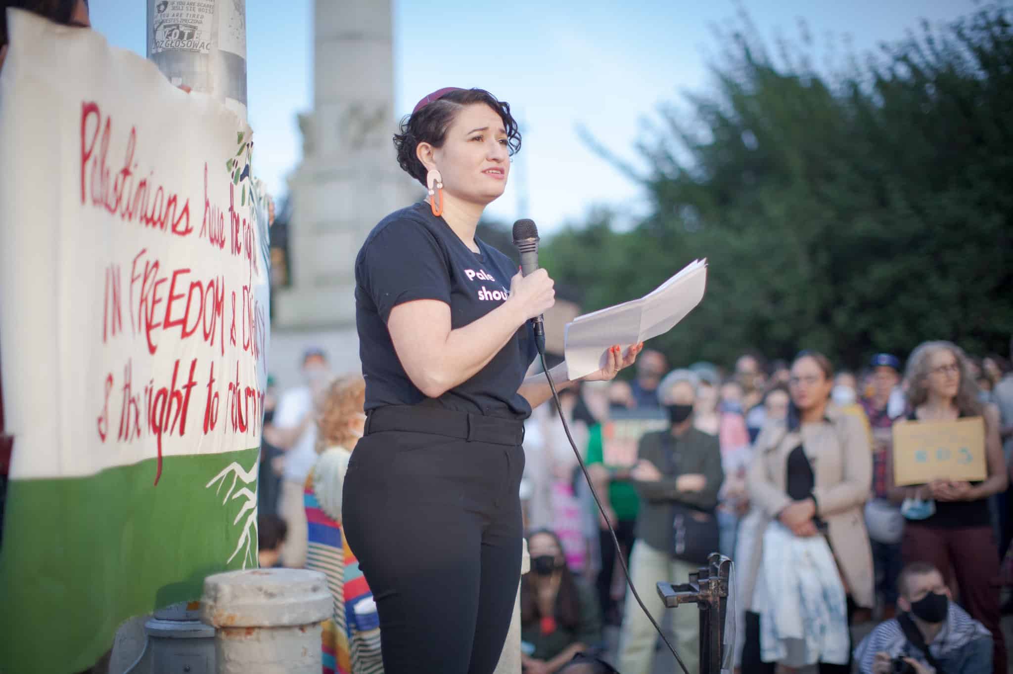 Rabbi Miriam Grossman speaks into a microphone at an anti-Israel demonstration, holding printed remarks while addressing a crowd gathered outdoors.