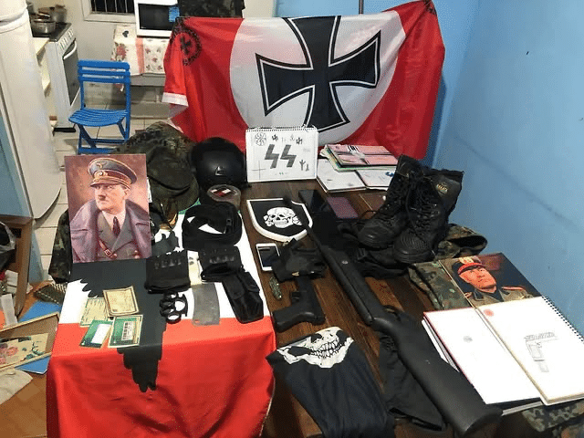 Table displaying neo-Nazi paraphernalia seized in Brazil, including Nazi and SS flags, Adolf Hitler portraits, military-style gear, helmets, boots, and weapons.