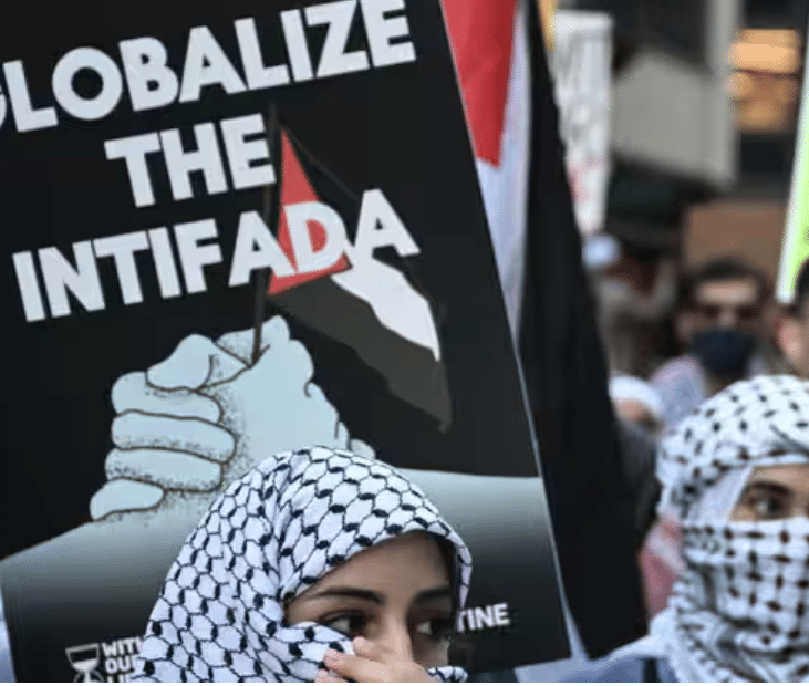 Protesters wearing keffiyehs stand in front of a sign reading “Globalize the Intifada” during a pro-Palestinian demonstration, highlighting the slogan’s role in contemporary anti-Israel activism.