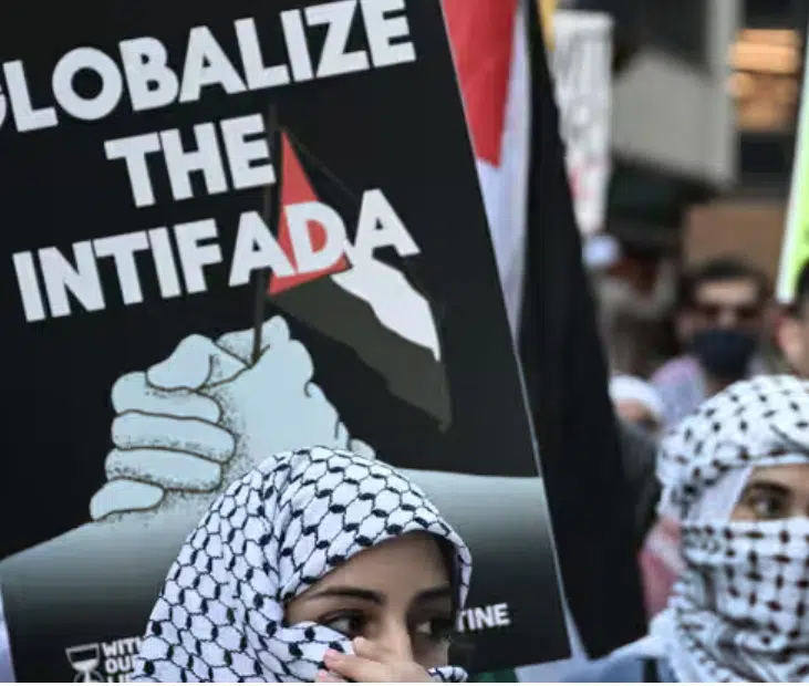 Protesters wearing keffiyehs stand in front of a sign reading “Globalize the Intifada” during a pro-Palestinian demonstration, highlighting the slogan’s role in contemporary anti-Israel activism.