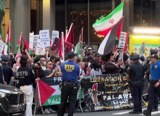 PAL-Awda demonstrators protest outside a Jerusalem investment expo in Manhattan while displaying Palestinian, Iranian, and PFLP flags behind NYPD officers and security barriers.