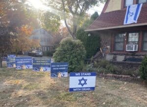 Israeli flag displayed on a house alongside lawn signs expressing support for Israel and solidarity with victims of the October 7 Hamas massacre.