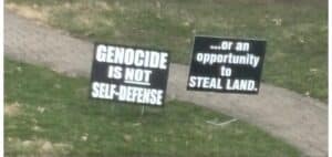Two lawn signs displayed on a residential property reading "Genocide Is Not Self-Defense" and "...or an opportunity to steal land."