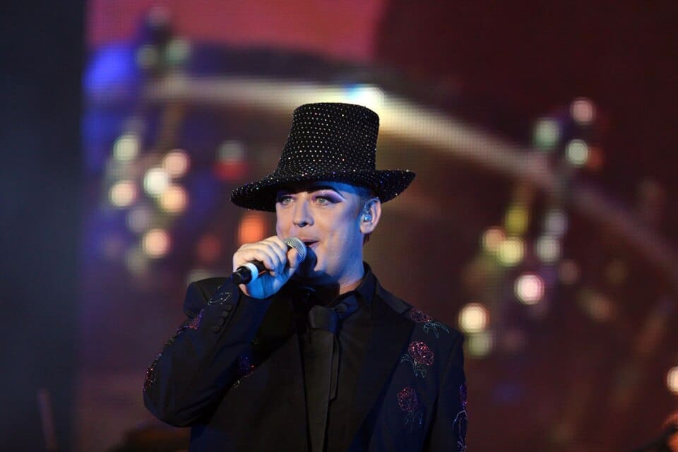 Boy George performs on stage during a live concert, wearing a black suit and wide-brimmed hat while singing into a microphone.