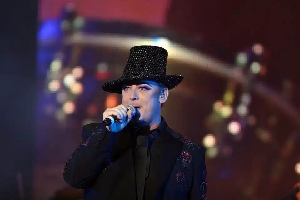 Boy George performs on stage during a live concert, wearing a black suit and wide-brimmed hat while singing into a microphone.