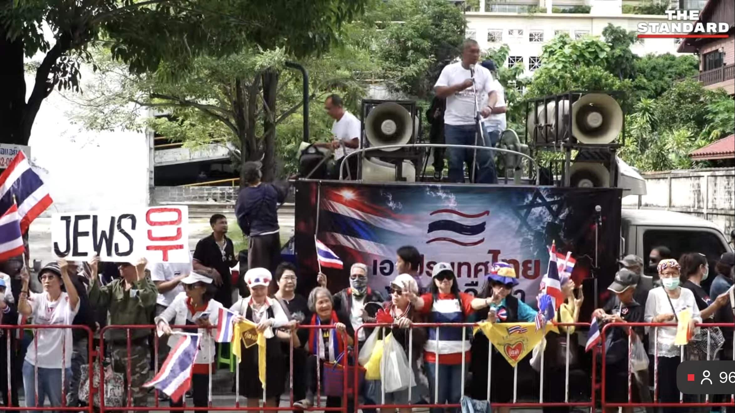 Thai protesters gather behind barriers in Bangkok as one demonstrator holds a sign reading “Jews out.”