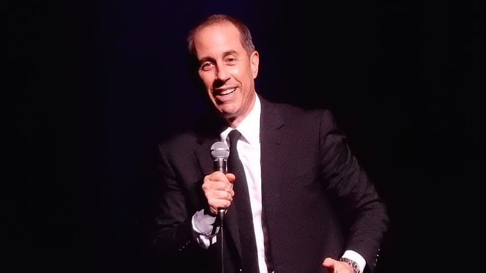 Jerry Seinfeld smiles while holding a microphone during a stand-up comedy performance.